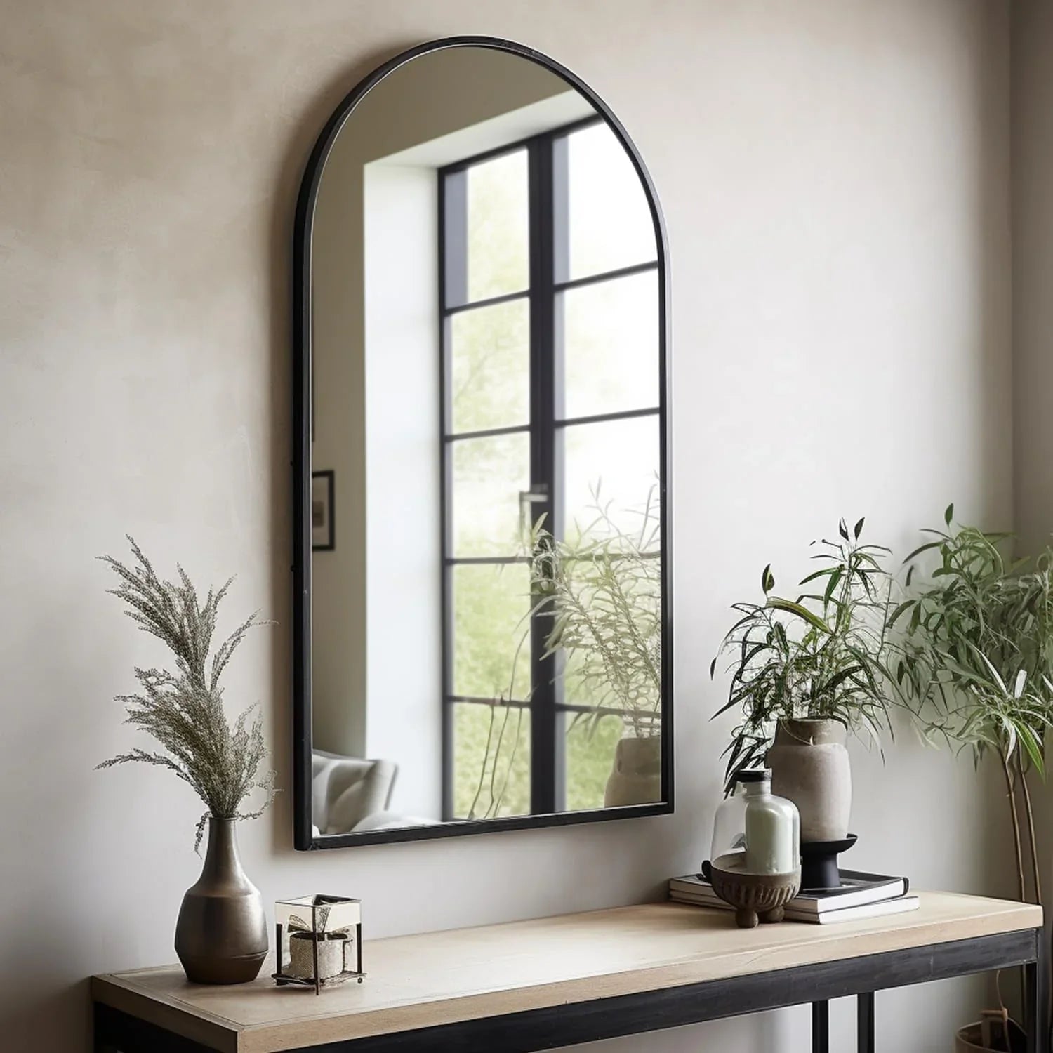 Black metal arched wall mirror mounted above wooden entryway console table with green plants.