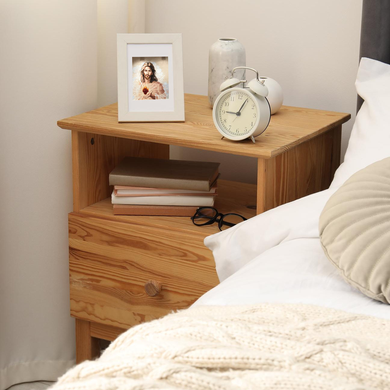 Natural pine wood nightstand with drawer, open shelf, retro alarm clock, and framed Jesus portrait.