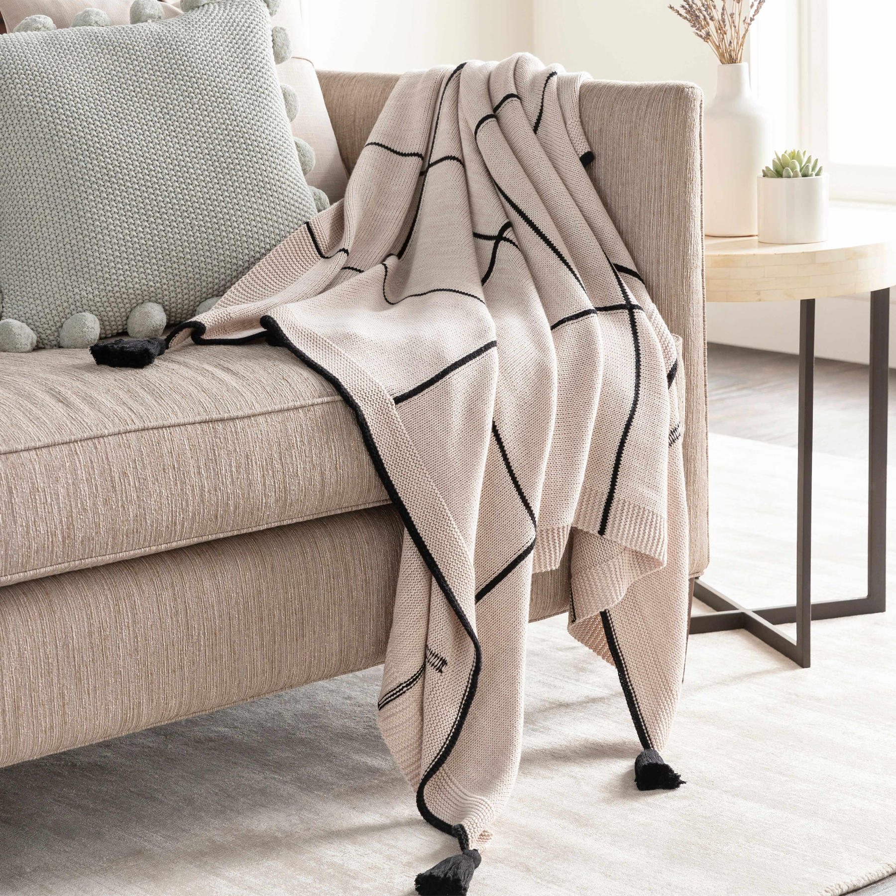 A cream knit throw blanket with black geometric lines and black pom-pom trim draped over a beige sofa in a bright living room setting.